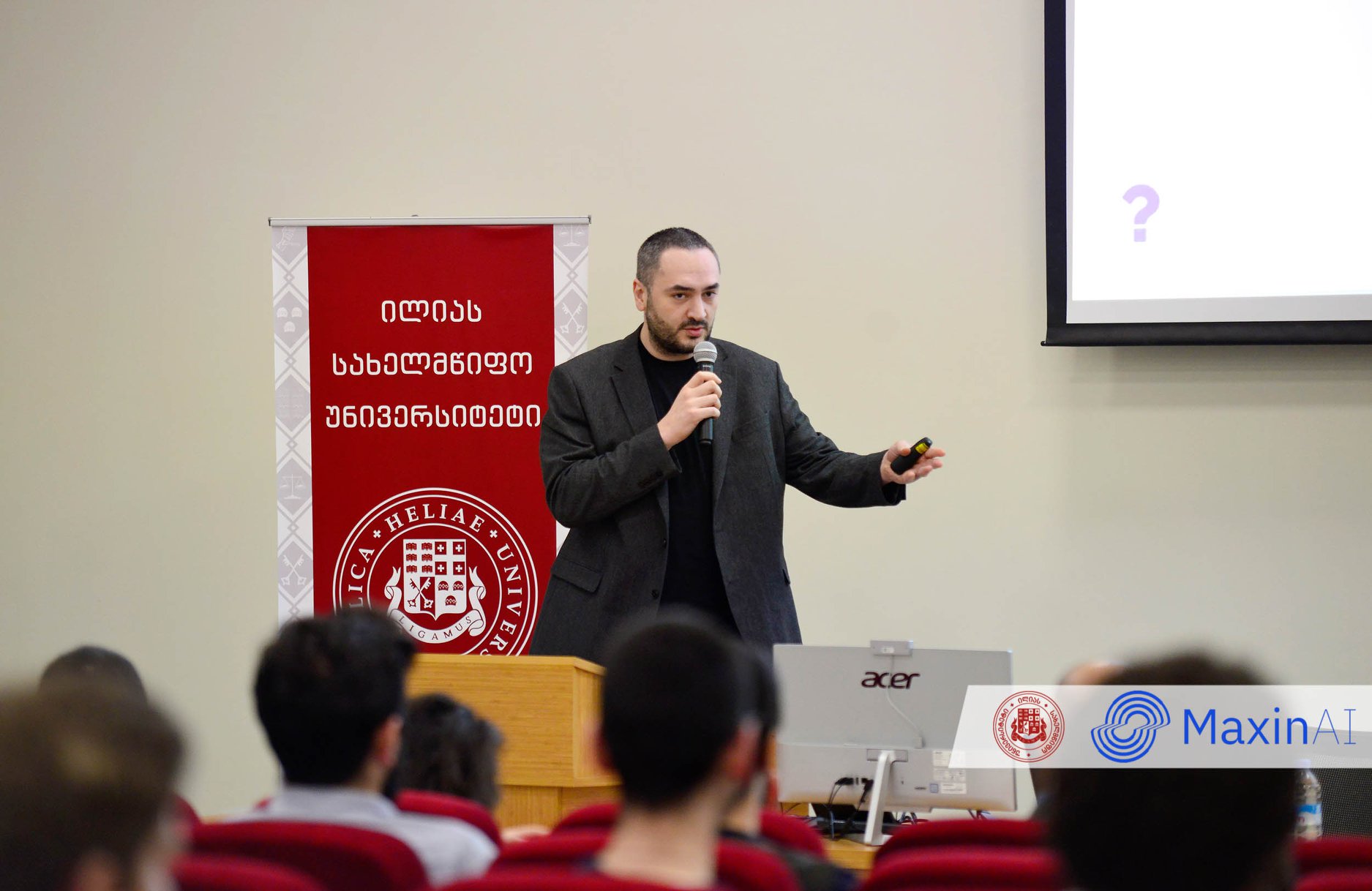 A speaker in a blazer holding a microphone presents at the front of a lecture hall, with a university banner behind and audience members in the foreground.