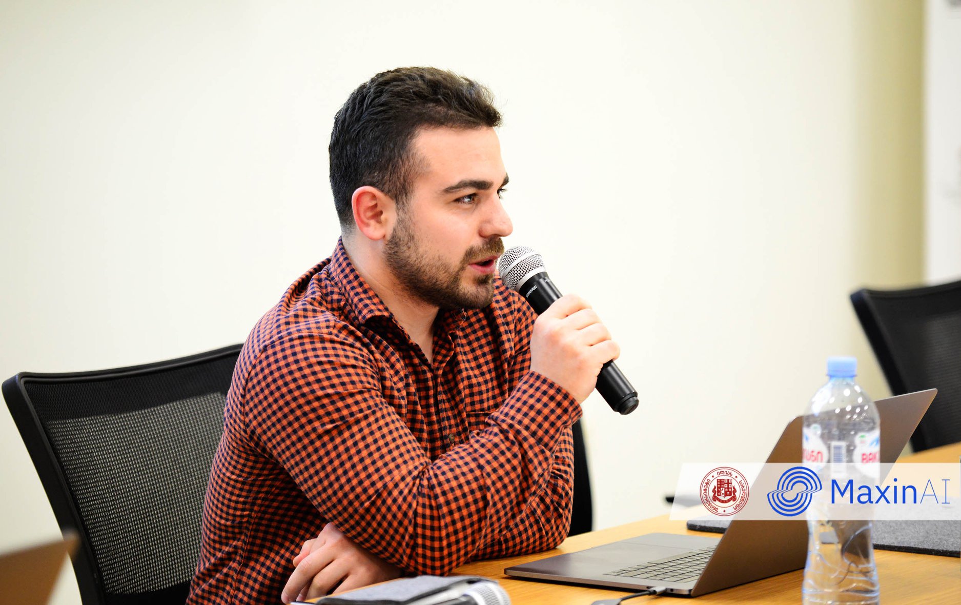 A young engineer in a checked shirt speaks into a microphone at a panel desk, with a laptop and water bottle in front.